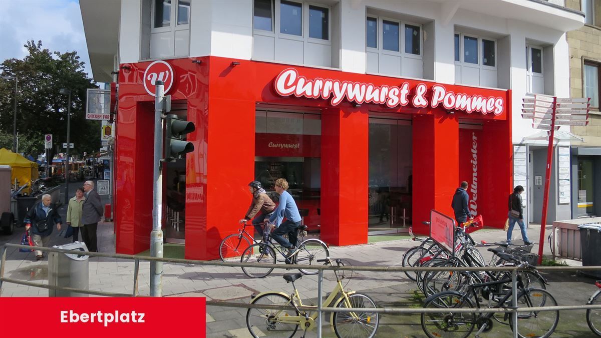 Leute auf Fahrrädern vor einem Gebäude mit der Aufschrift „Currywurst & Pommes“