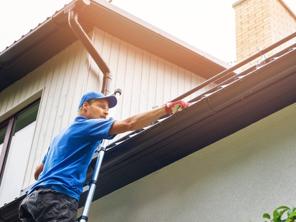 Un hombre está de pie en una escalera arreglando una canaleta en el costado de una casa.