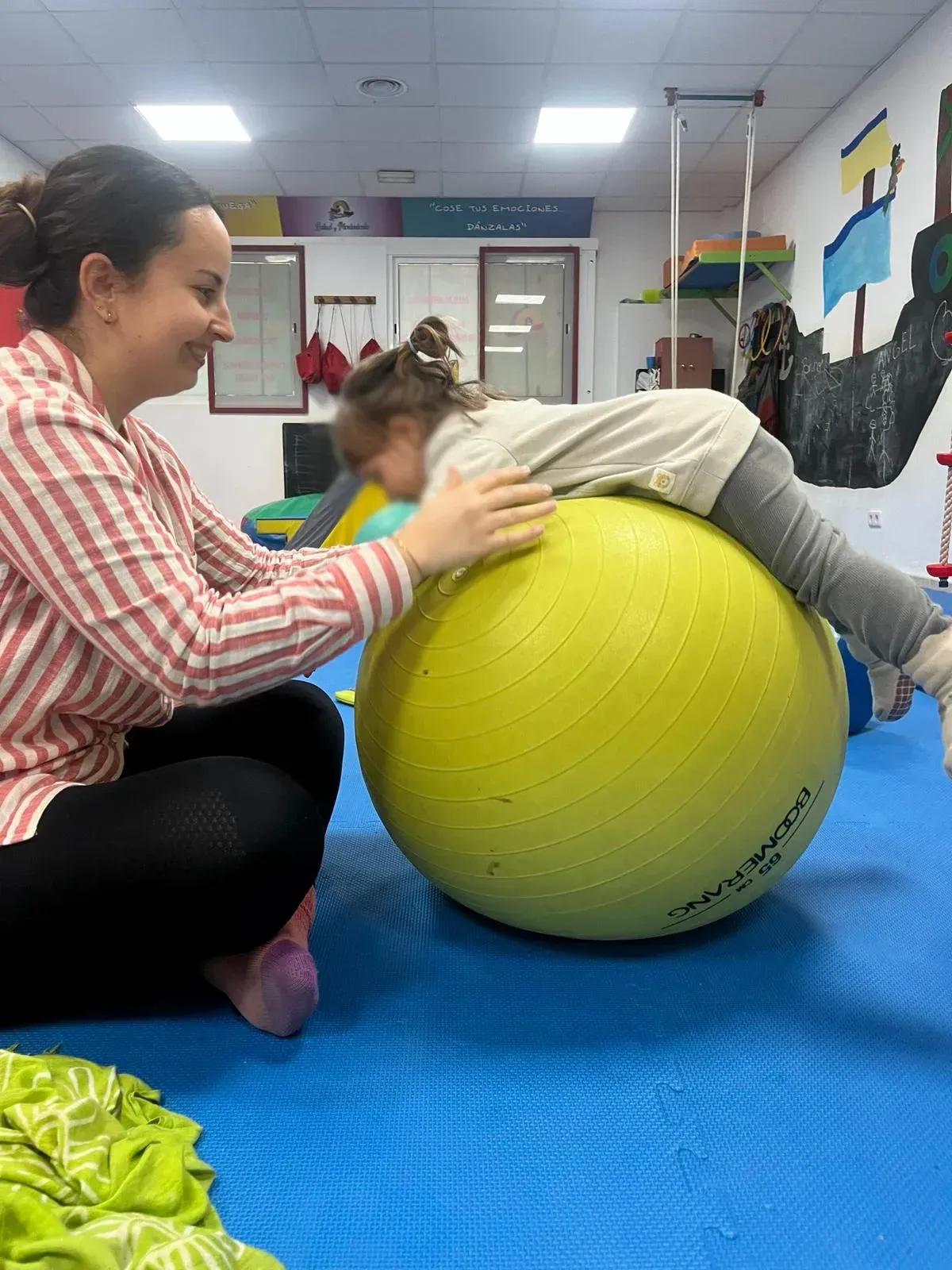 Una mujer está colocando a un niño sobre una pelota de ejercicio amarilla.
