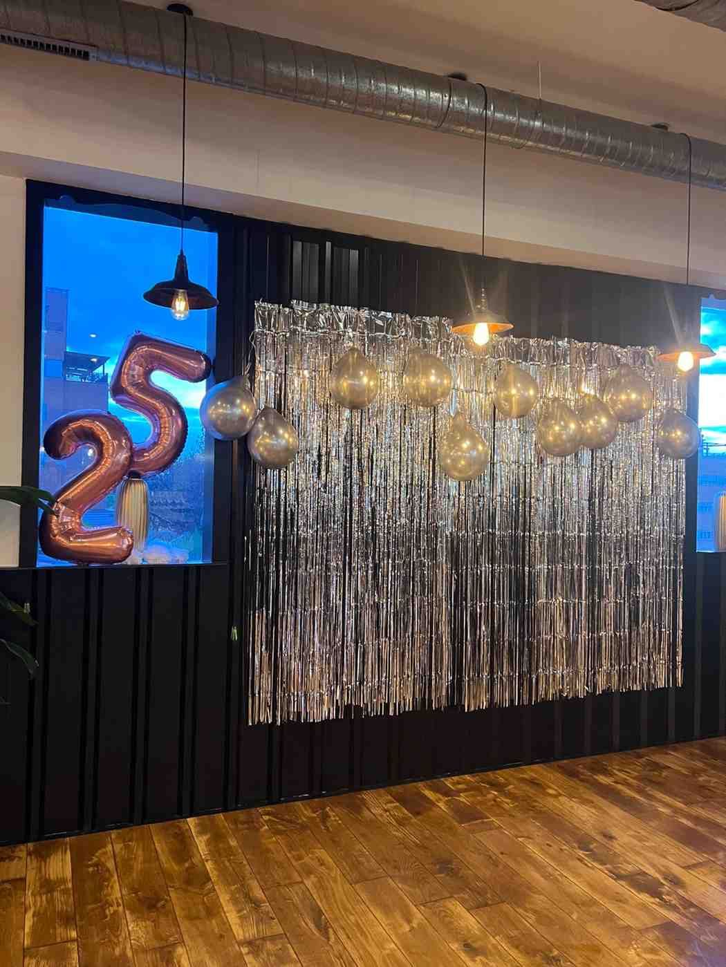 Fondo de fiesta con cortina plateada de oropel, globos y decoraciones con globos de 25 pulgadas. Suelo de madera y ventanal.