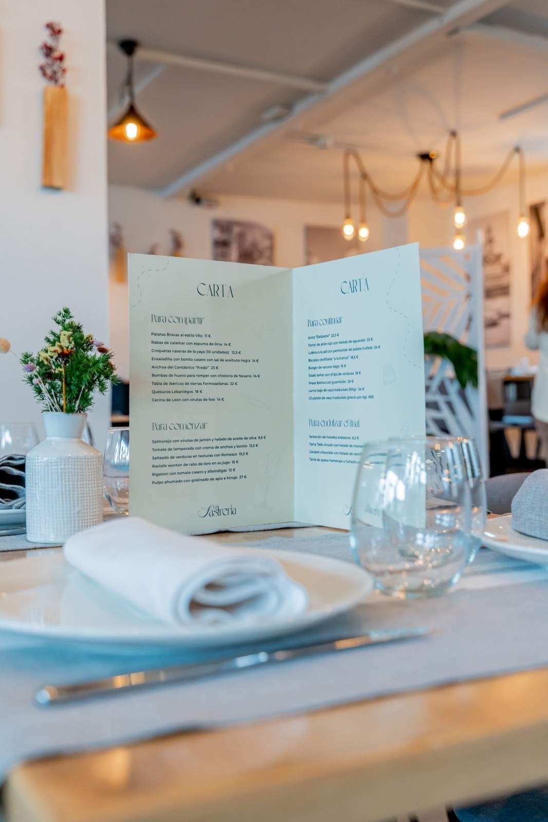 Una mesa de restaurante preparada con menú, platos, cristalería y un pequeño jarrón con flores.