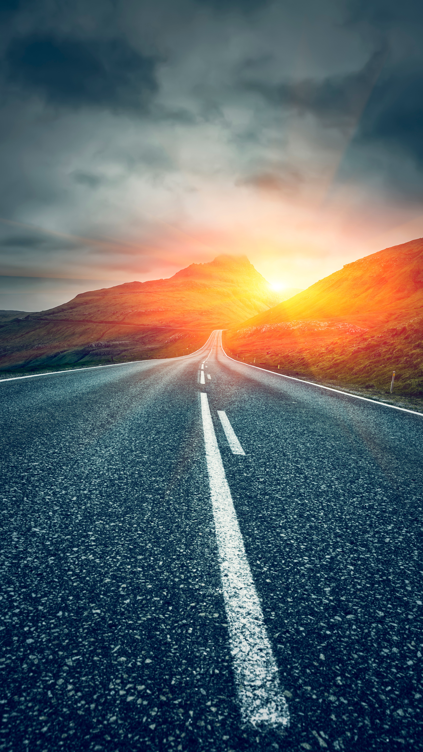 Route menant vers un coucher de soleil éclatant entre les collines, sous un ciel nuageux.