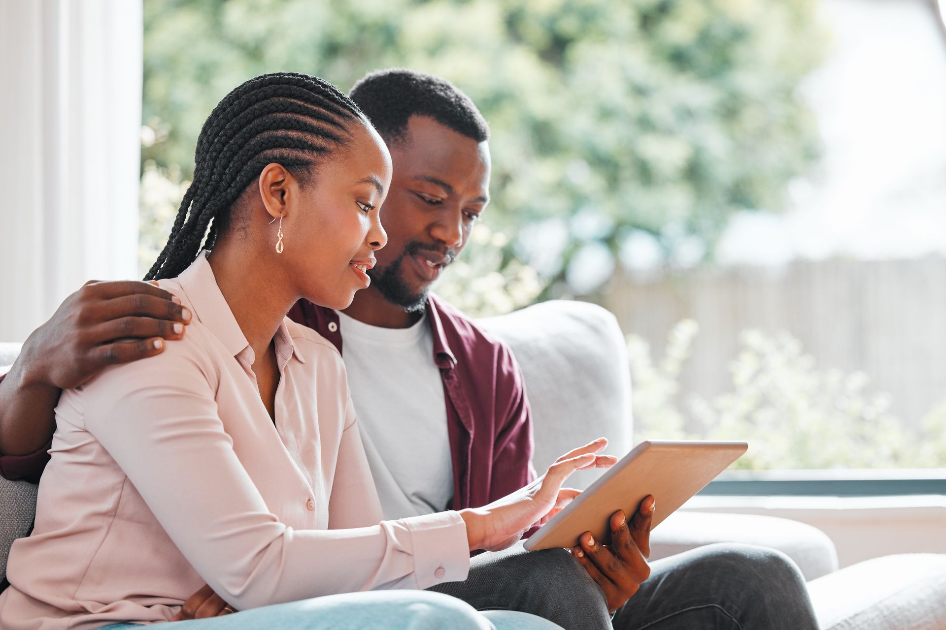 Un couple regarde une tablette sur un canapé, à l'intérieur près d'une fenêtre ; la femme montre du doigt.