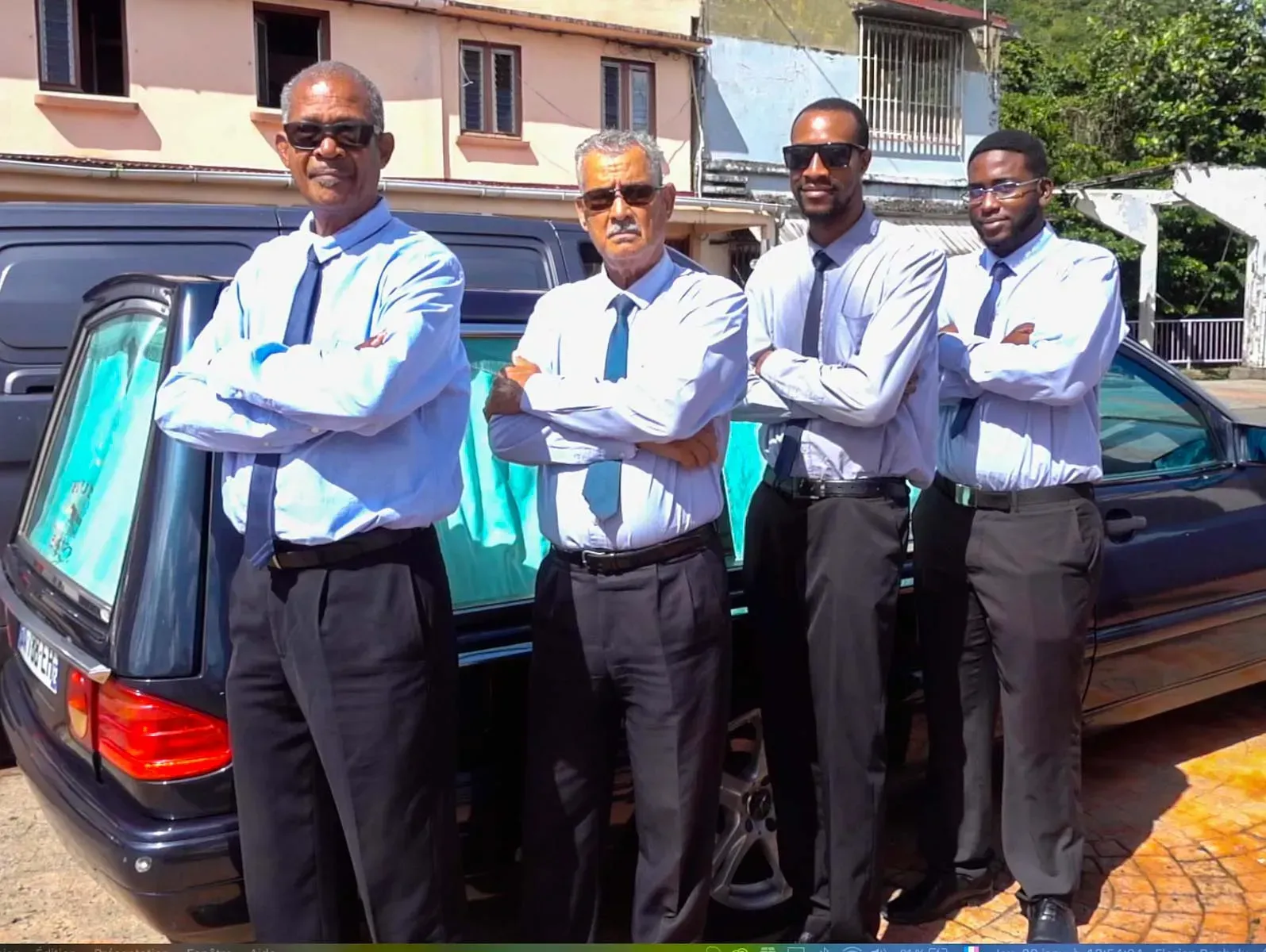 Quatre conseillers funéraires en chemises bleu clair et cravates se tiennent près d'un corbillard, les bras croisés.