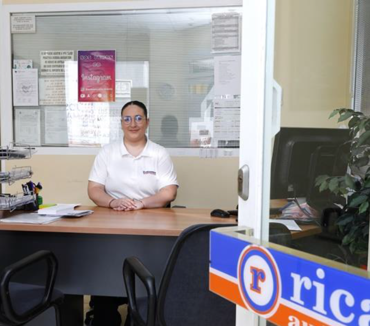 Mujer en el escritorio sonriendo, detrás de una computadora, fondo de oficina con logotipos.
