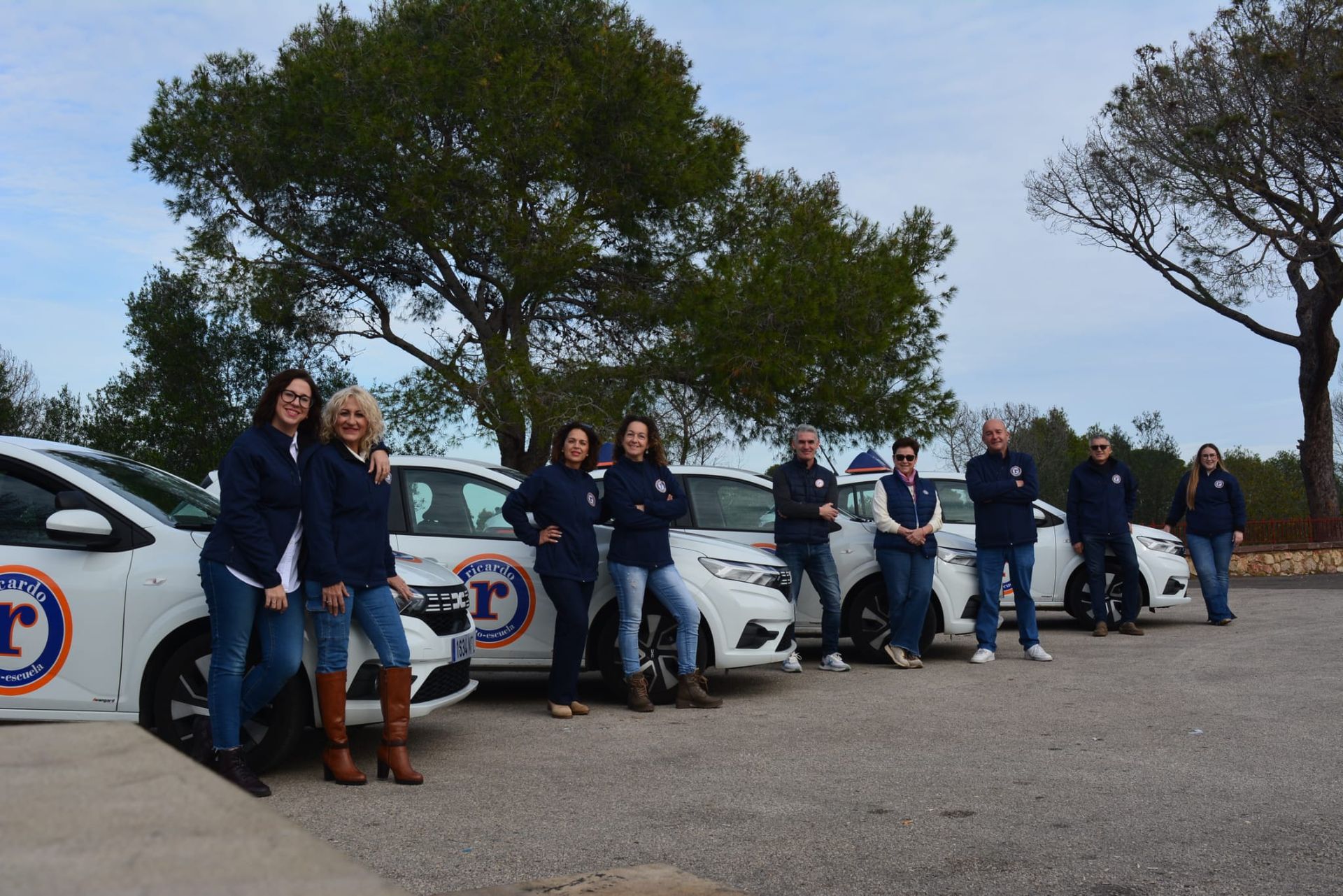 Un grupo de personas con camisas azules y pantalones vaqueros se encuentran al lado de coches blancos de marca al aire libre.