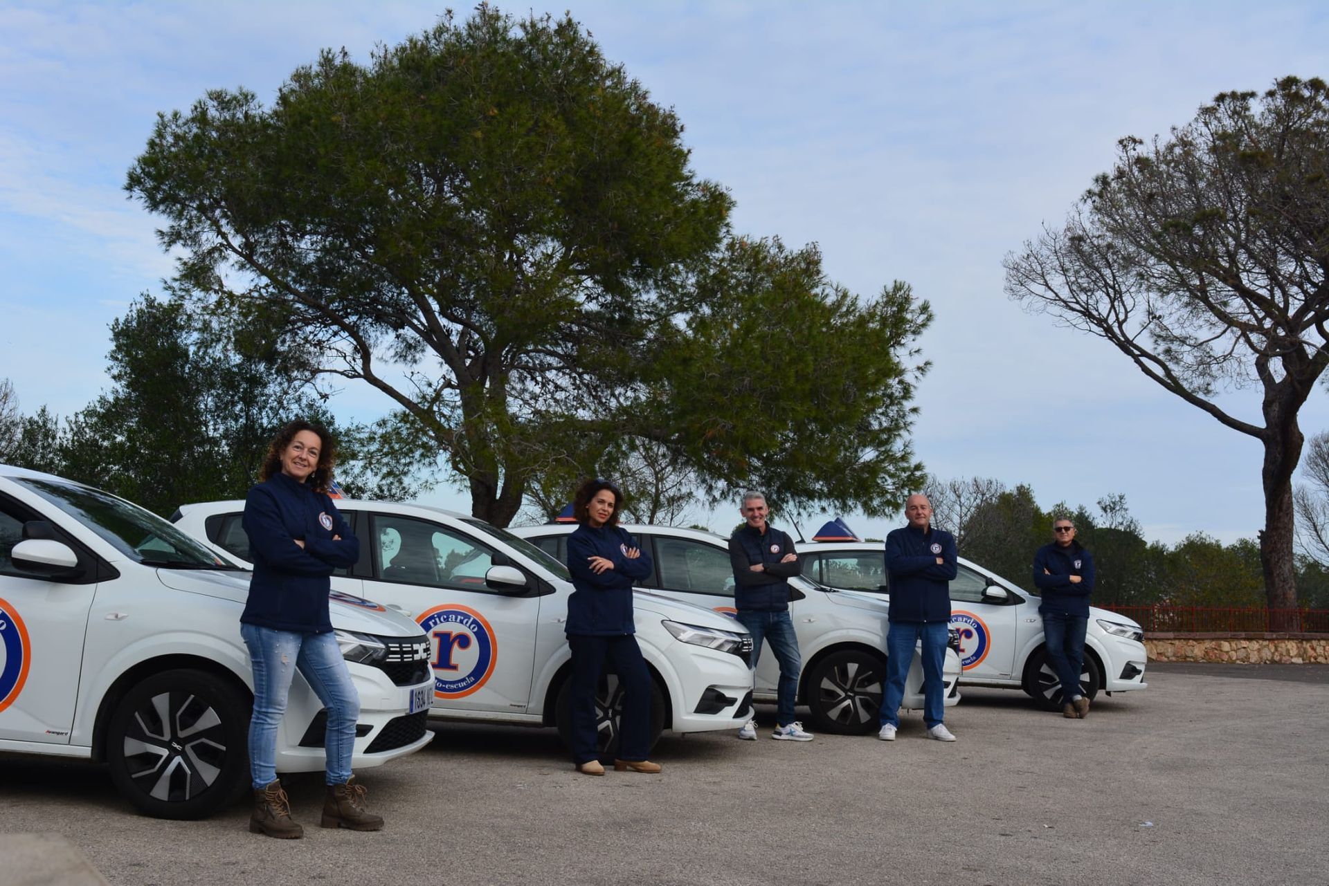Cinco personas están de pie junto a coches blancos con logotipos. Están afuera, con árboles al fondo.