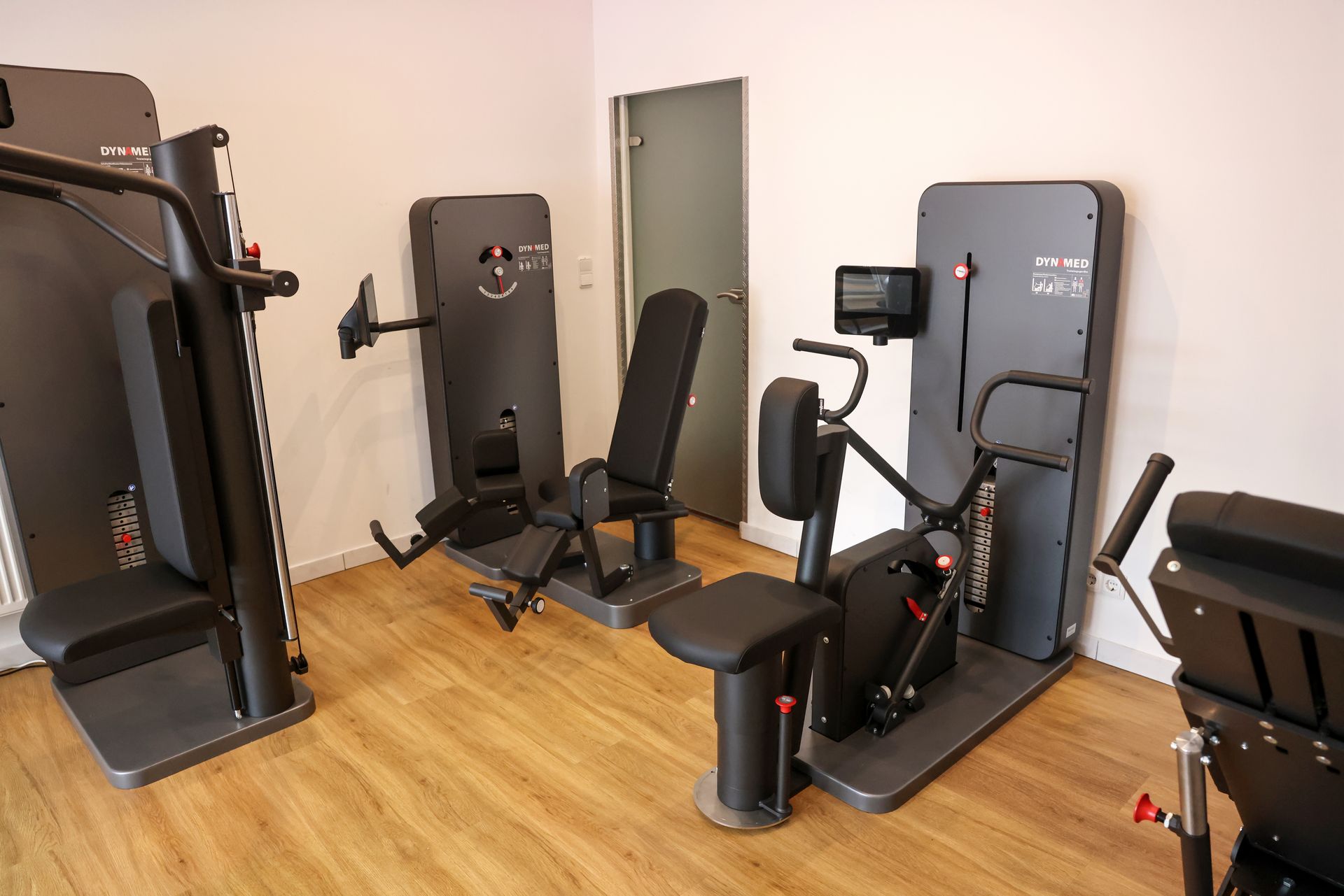 Mehrere moderne Trainingsgeräte stehen in einem hellen Fitness- und Therapieraum mit Holzfußboden.