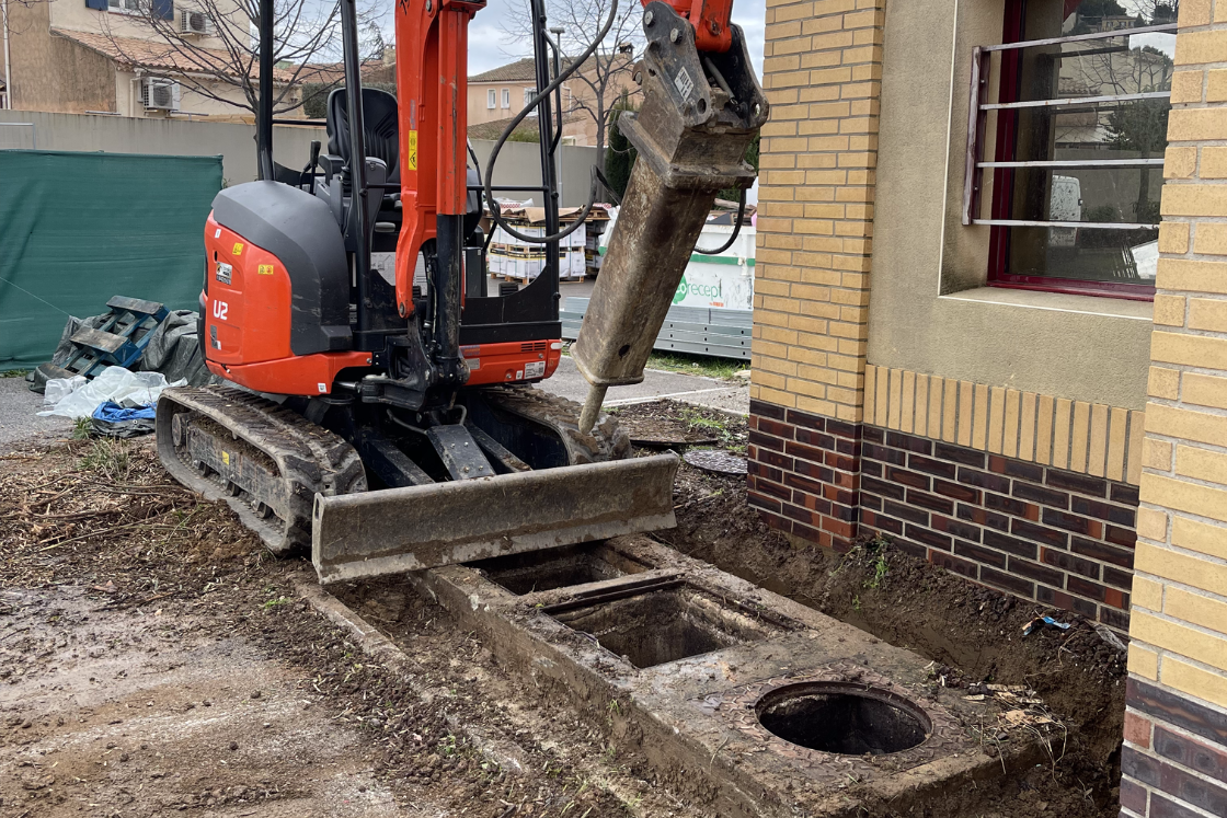 Une excavatrice creuse un trou dans le sol devant un bâtiment en briques.