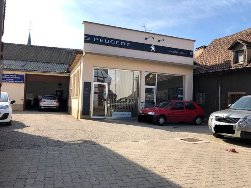 Concessionnaire automobile Peugeot avec voitures garées à l'extérieur, voiture rouge devant, journée ensoleillée.