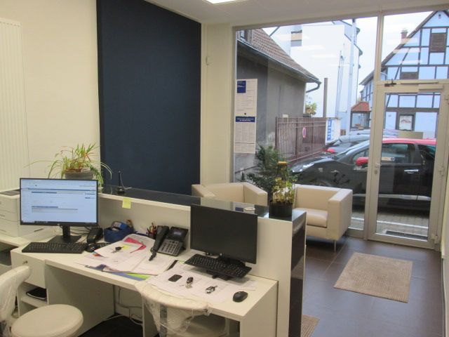 Bureau d'accueil dans un cabinet médical avec un ordinateur, un téléphone et une salle d'attente visibles à travers une porte vitrée.