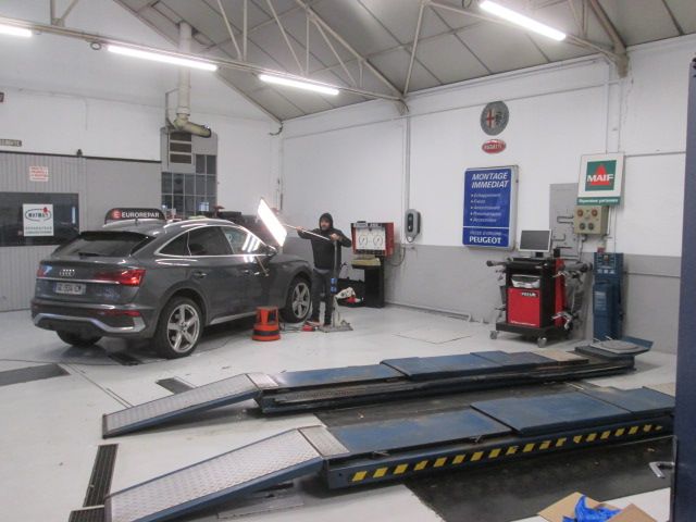 Voiture en cours d'entretien dans un garage, avec une personne travaillant dessus. Des rampes, des outils et du matériel sont visibles.