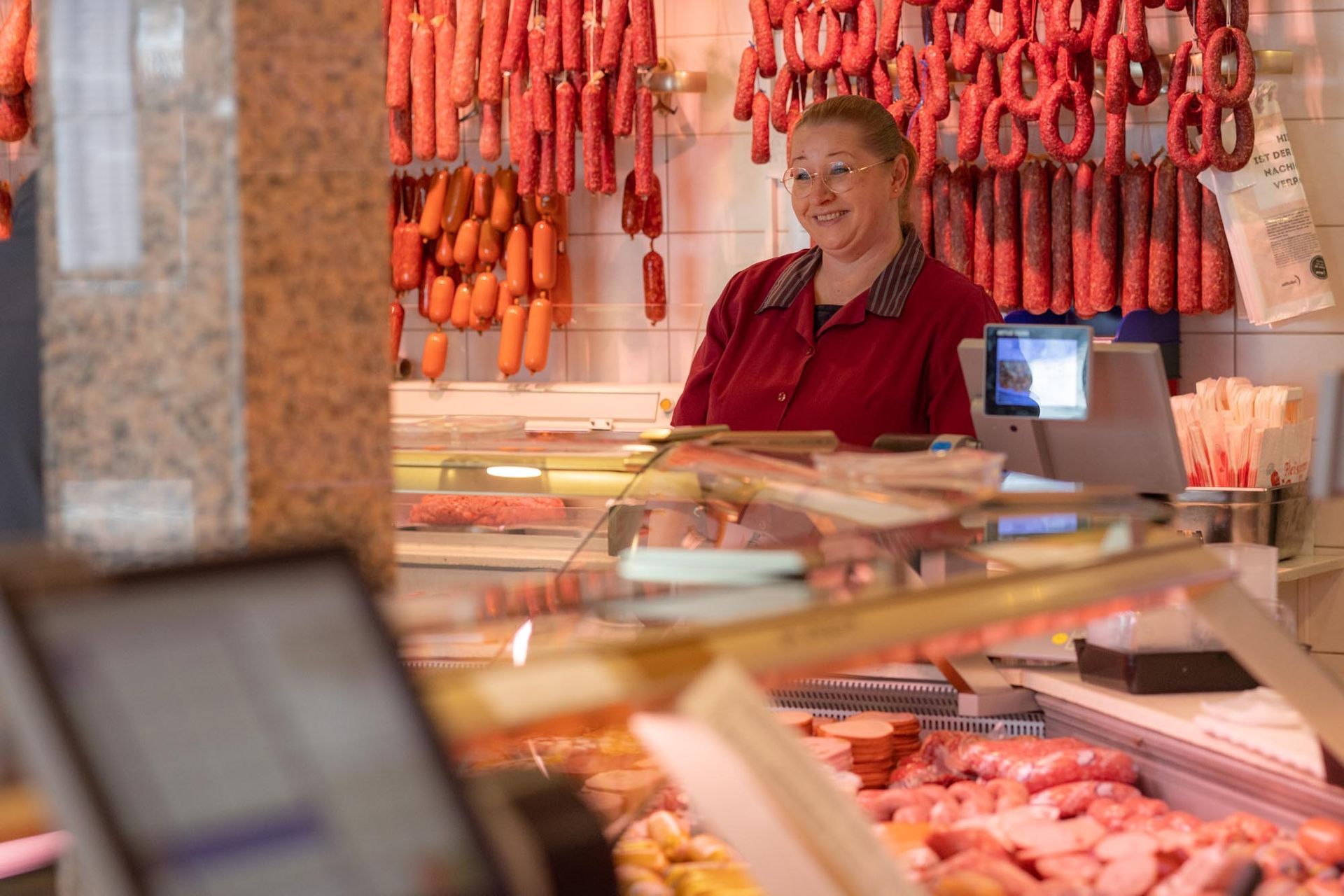 Ein Verkäufer lächelt hinter einem Marktstand mit hängenden Würsten und Frischfleischauslagen.