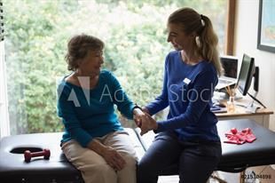 Eine ältere Frau sitzt auf einem Bett und hält die Hand einer Krankenschwester.