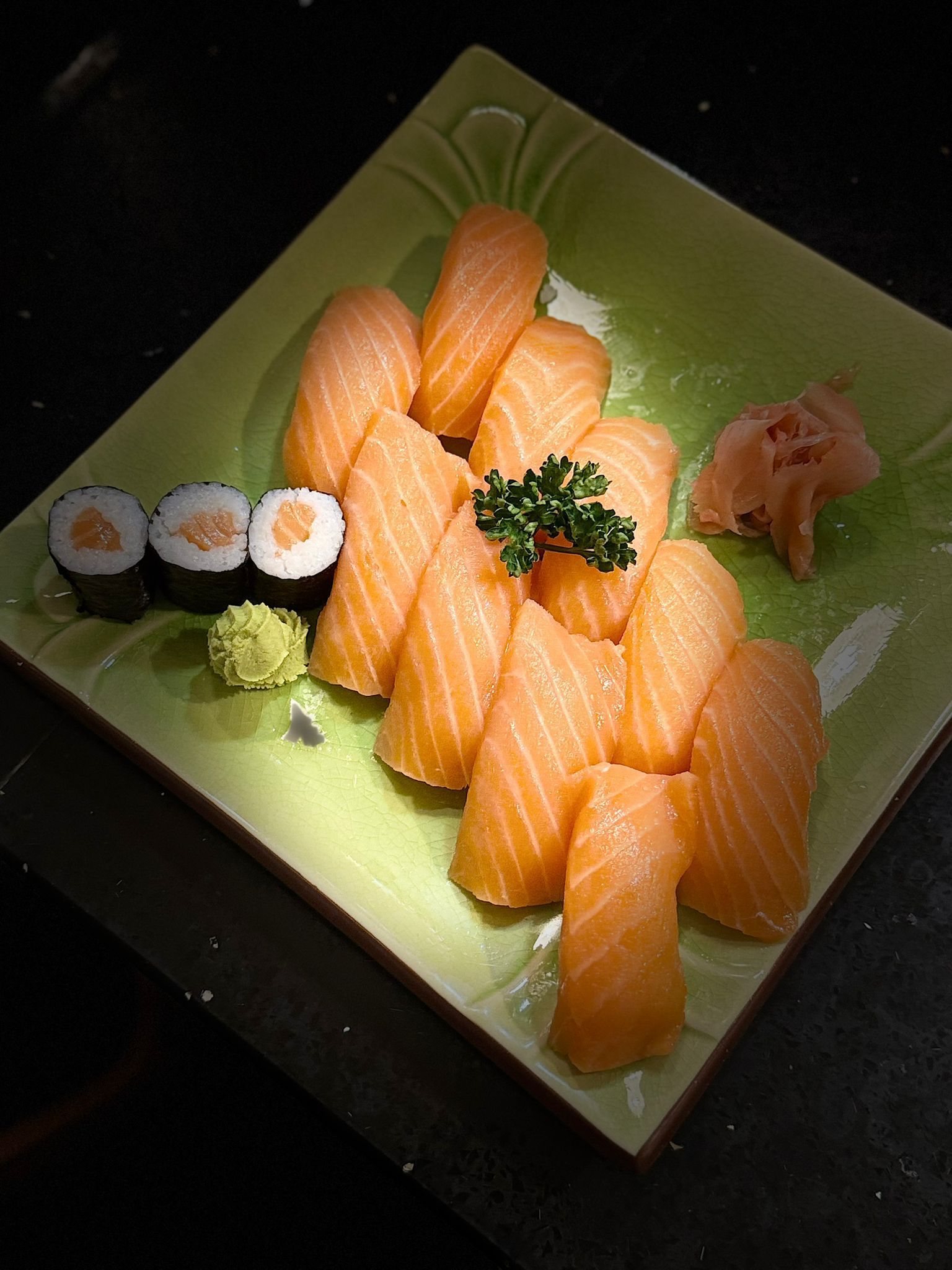 Ein Teller mit Lachs-Sushi, Sushi-Rollen, Wasabi und Ingwer auf einem grünen, quadratischen Teller.