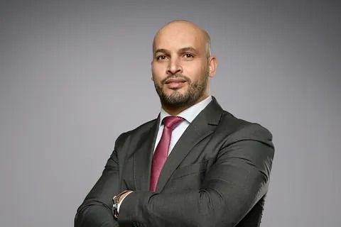 Hombre de traje con los brazos cruzados, sonriendo. Fondo gris.