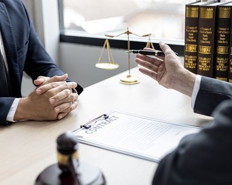 Consulta legal: una persona con traje se sienta frente a otra con toga. Sobre un escritorio hay balanzas de la justicia, libros de leyes y un contrato.