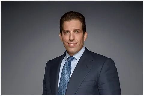 Hombre con traje y corbata azules sonriendo sobre un fondo gris.