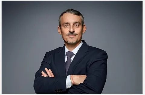 Hombre de traje con los brazos cruzados, sonriendo levemente, sobre un fondo gris.