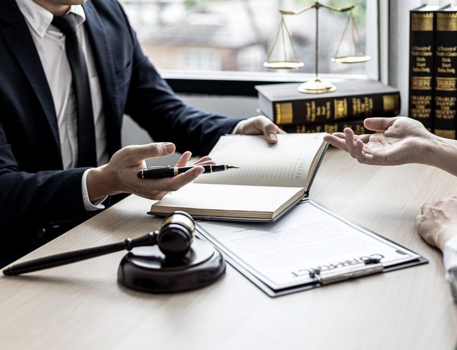 Abogado consultando con un cliente, señalando un bloc de notas. Presencia de mazo, balanza de la justicia y libros de derecho.