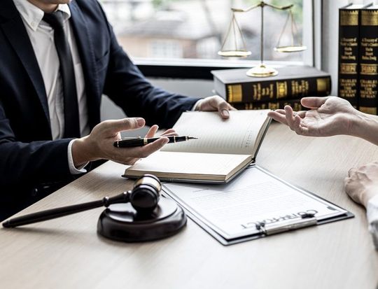 Abogado consultando con un cliente, señalando un bloc de notas. Presencia de mazo, balanza de la justicia y libros de derecho.