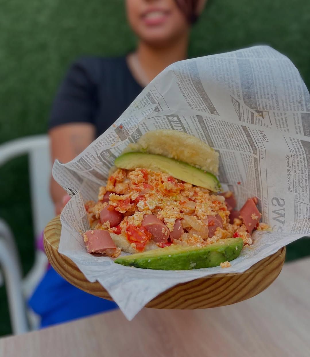 Persona sosteniendo una arepa venezolana rellena de huevos revueltos, tomates y salchichas.