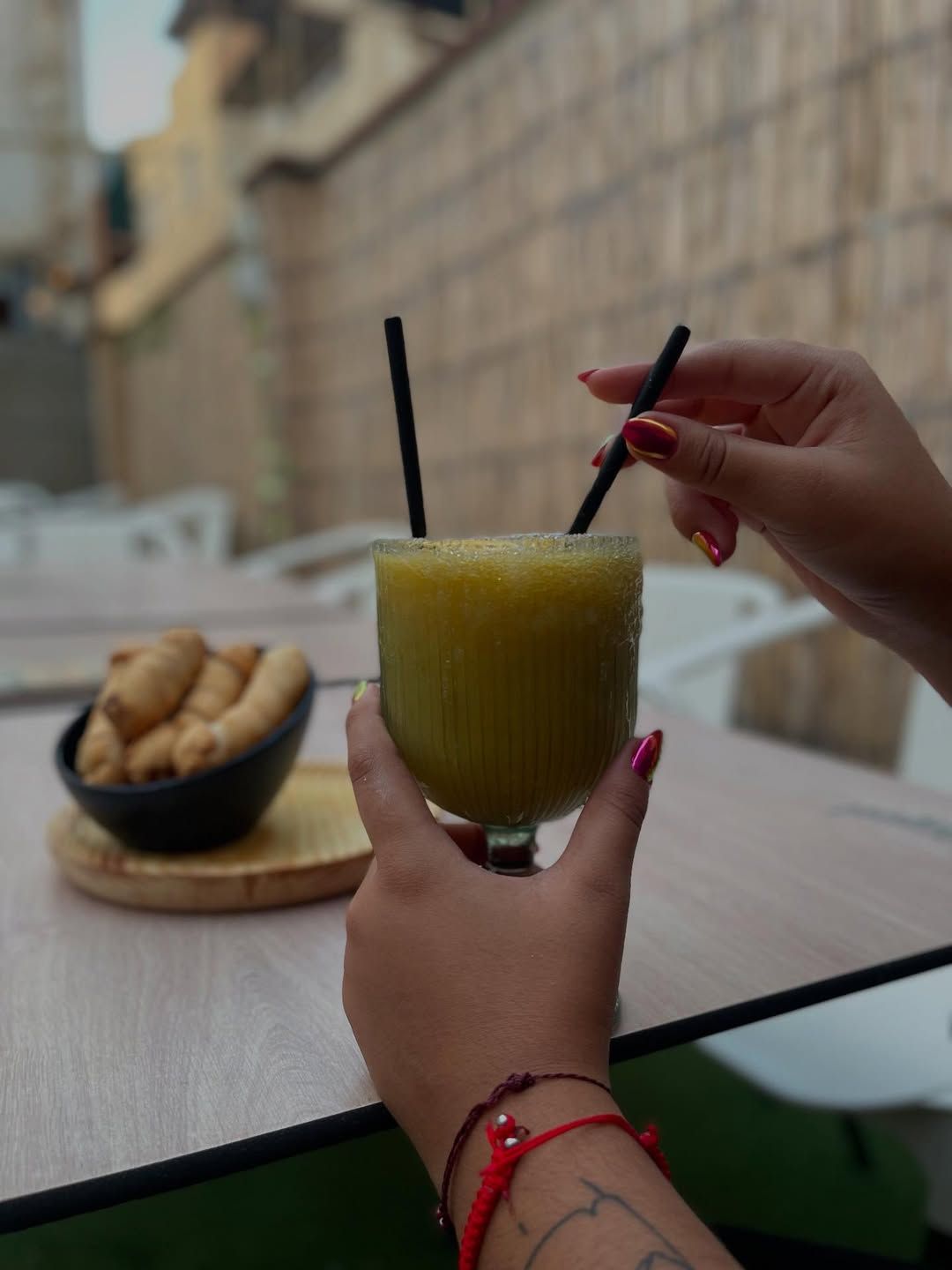 Una persona sostiene coctel con dos pajitas, cerca de un recipiente para bocadillos y asientos al aire libre.