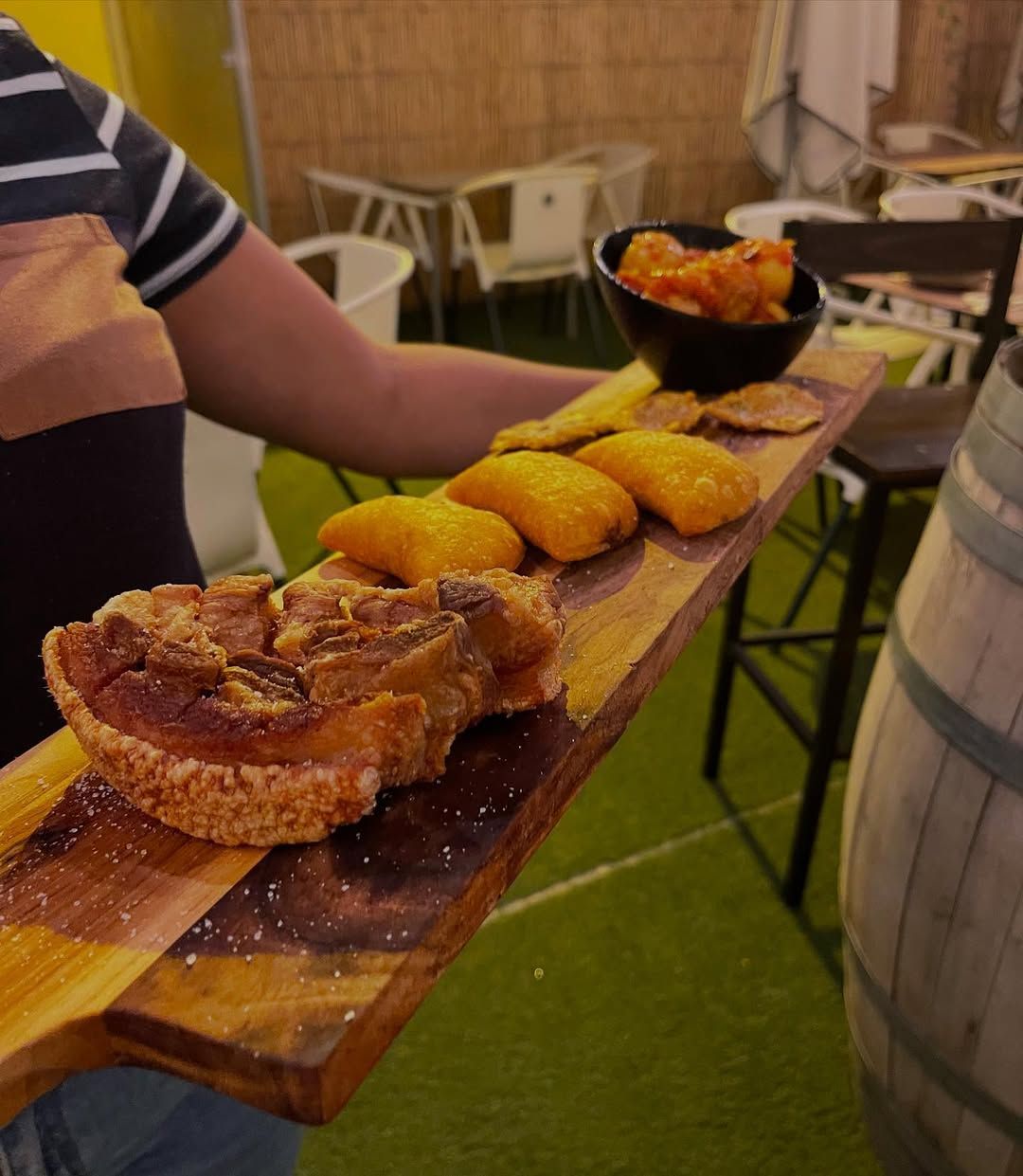 Persona sosteniendo una tabla de madera con cerdo frito, empanadas y papas.