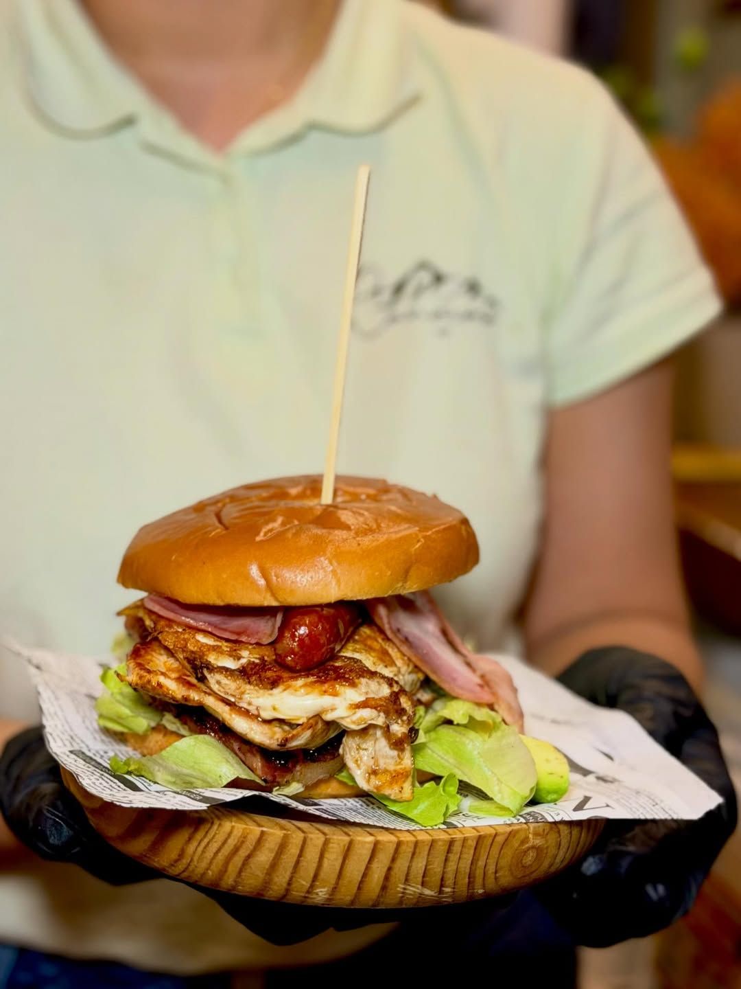 Una persona sostiene una hamburguesa grande en un plato de madera. La hamburguesa tiene varias capas y está sujeta con un palillo.