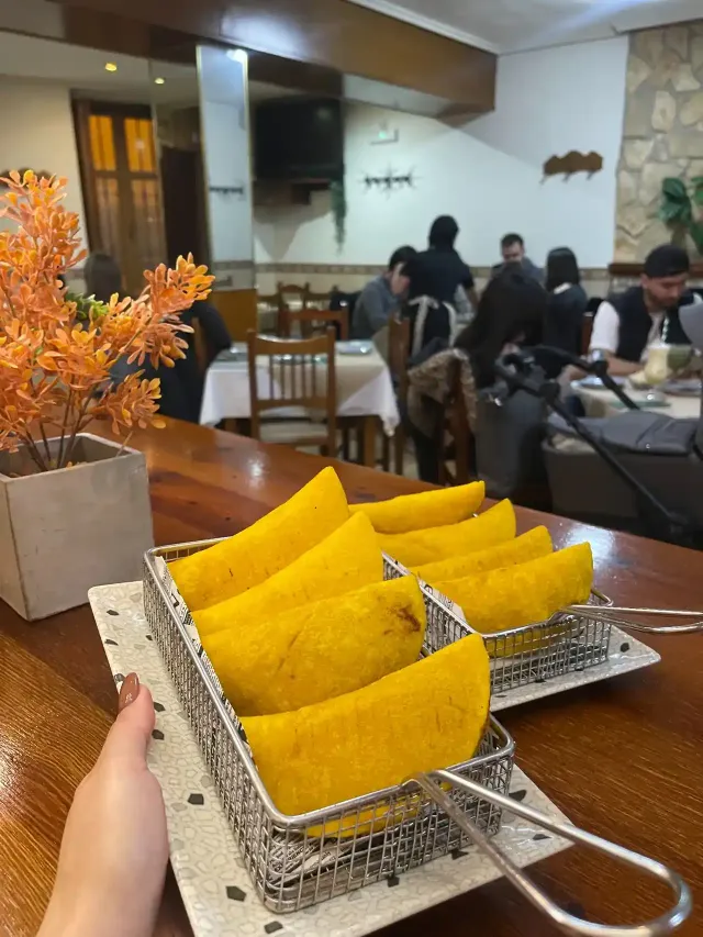 Dos cestas de empanadas amarillas sobre una mesa de madera en un restaurante. Personas sentadas al fondo.