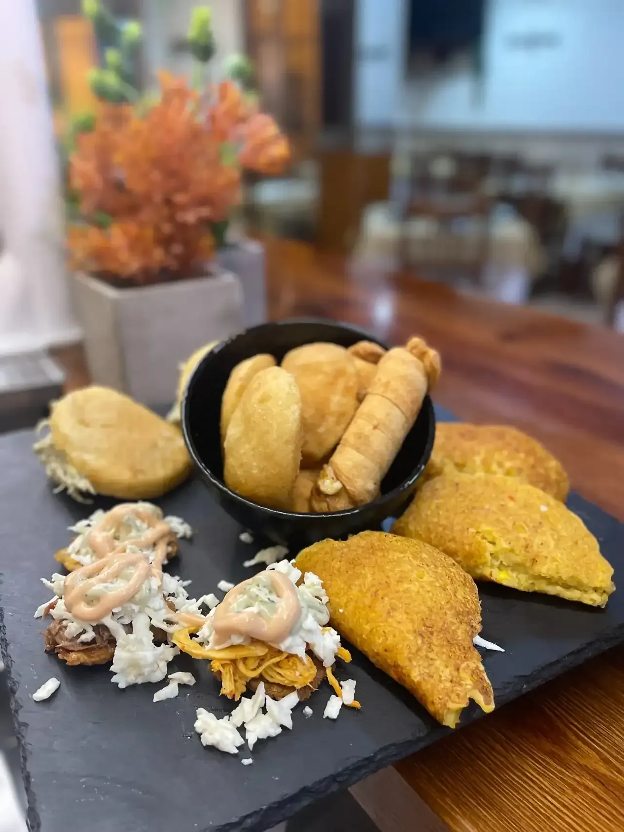 Surtido de bocadillos latinoamericanos fritos sobre pizarra negra, incluyendo empanadas, arepas y tequeños.