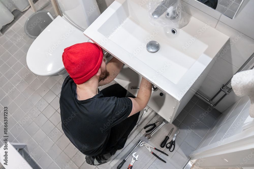 Eine Person mit roter Mütze arbeitet an der Sanitärinstallation unter einem weissen Waschbecken im Badezimmer; Werkzeuge liegen daneben auf dem gefliesten Boden.