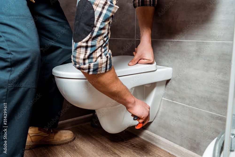 Ein Klempner in Arbeitskleidung installiert eine weisse, wandhängende Toilette in einem Badezimmer mit grau gefliesten Wänden.