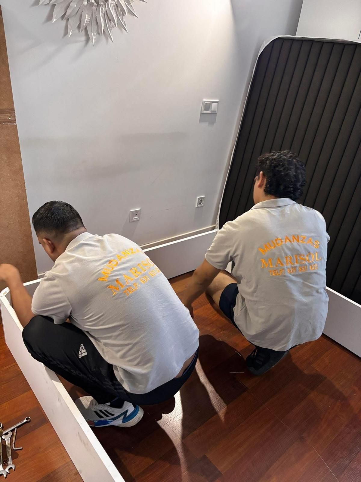 Dos trabajadores con camisas grises están agachados junto a una pared en el rellano de una escalera de madera, inspeccionando algo cerca del suelo.