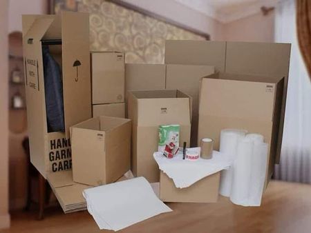 Cajas de cartón para mudanzas y material de embalaje apilados en una habitación con un sofá y una cortina.