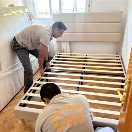 Dos trabajadores ensamblan la estructura de una cama de madera en un dormitorio con cabecero tapizado en blanco.