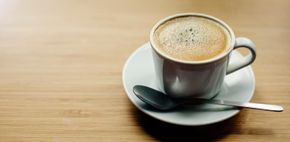 Taza de café sobre un platillo con una cuchara sobre una mesa de madera, con espuma encima.