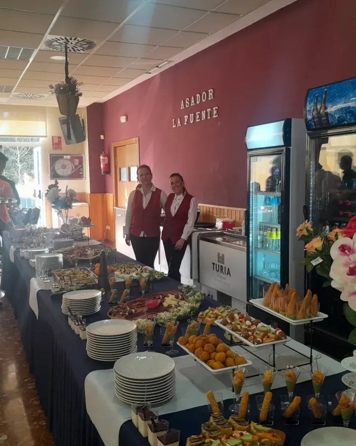 Mesa de buffet con pasteles y fruta en un restaurante, con dos empleados de pie detrás.