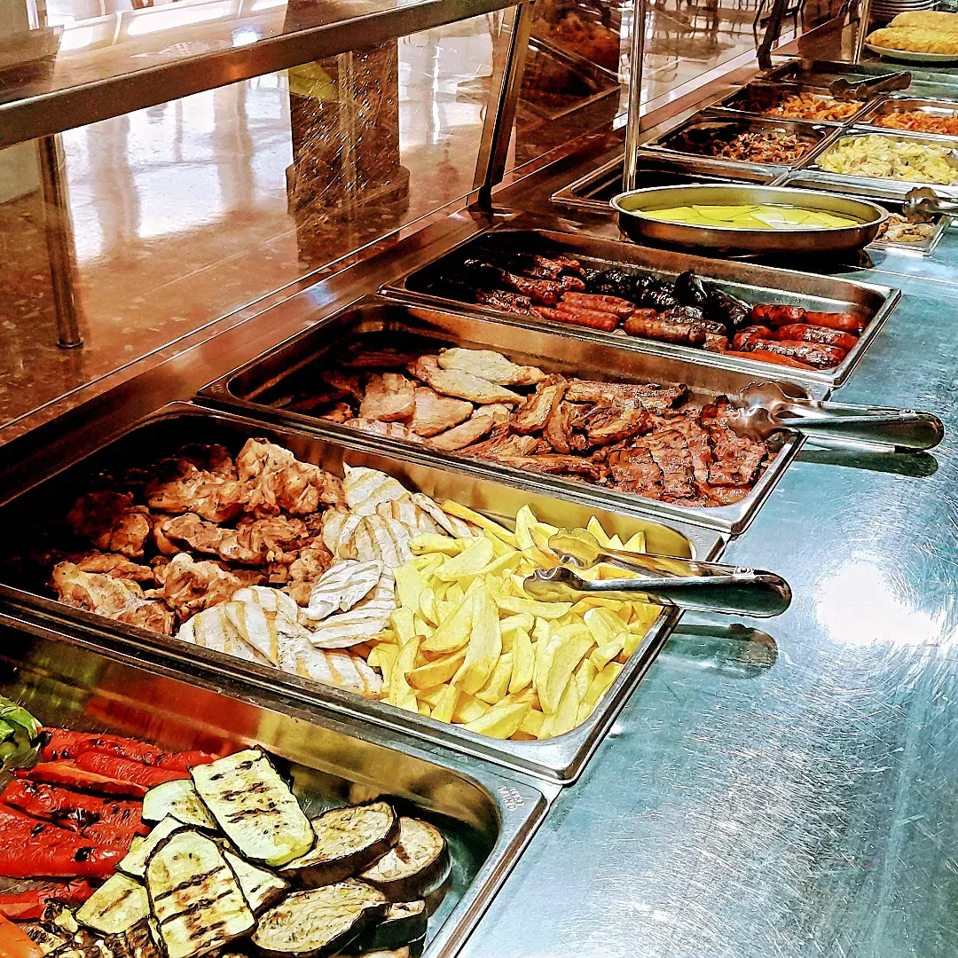 Línea de buffet con bandejas de carnes a la parrilla variadas, papas fritas, verduras y arroz sobre un mostrador brillante.