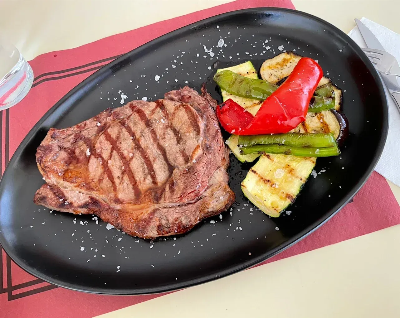 Filete a la parrilla en un plato ovalado negro con verduras asadas, sobre una mesa decorada en rojo y blanco.
