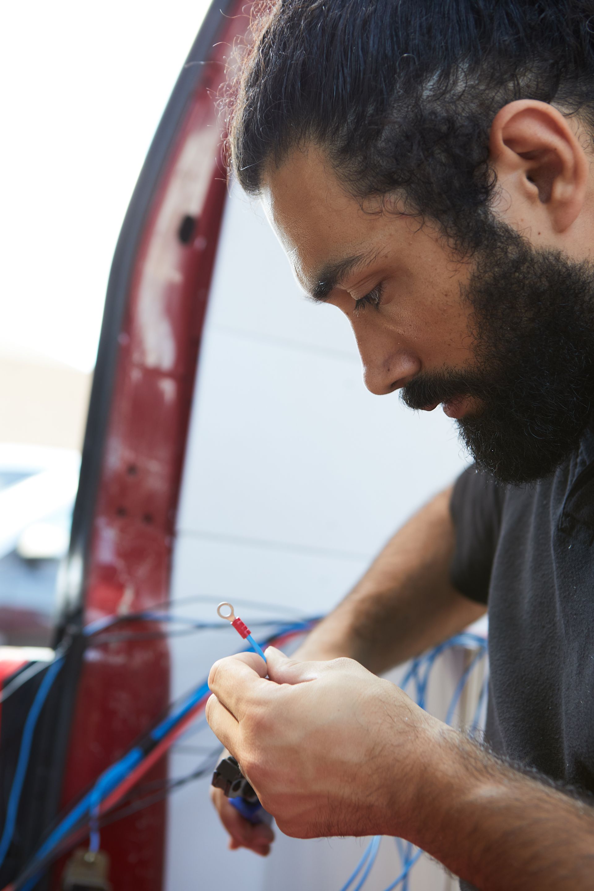 Homme barbu, concentré sur le câblage électrique près d'une porte de véhicule rouge ouverte.