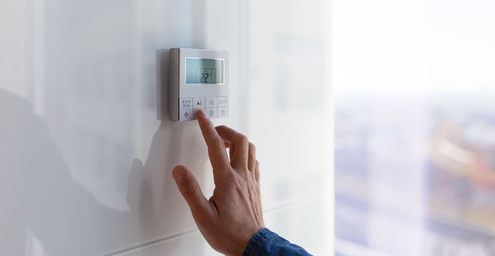 Main réglant un thermostat sur un mur blanc à côté d'une fenêtre.