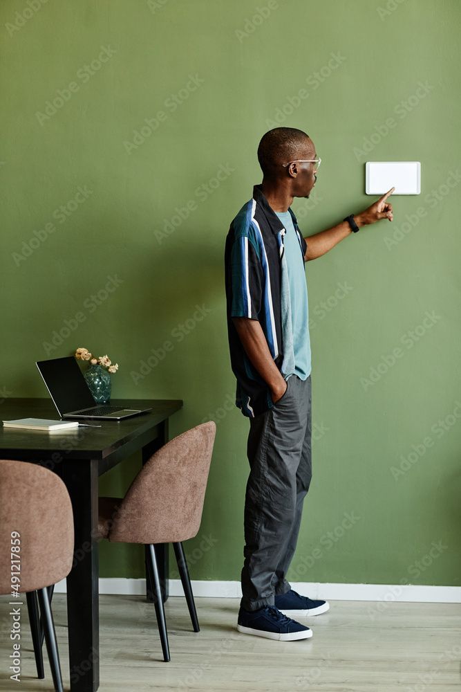 Un homme noir portant des lunettes pointe un thermostat sur un mur vert près d'une table avec un ordinateur portable.