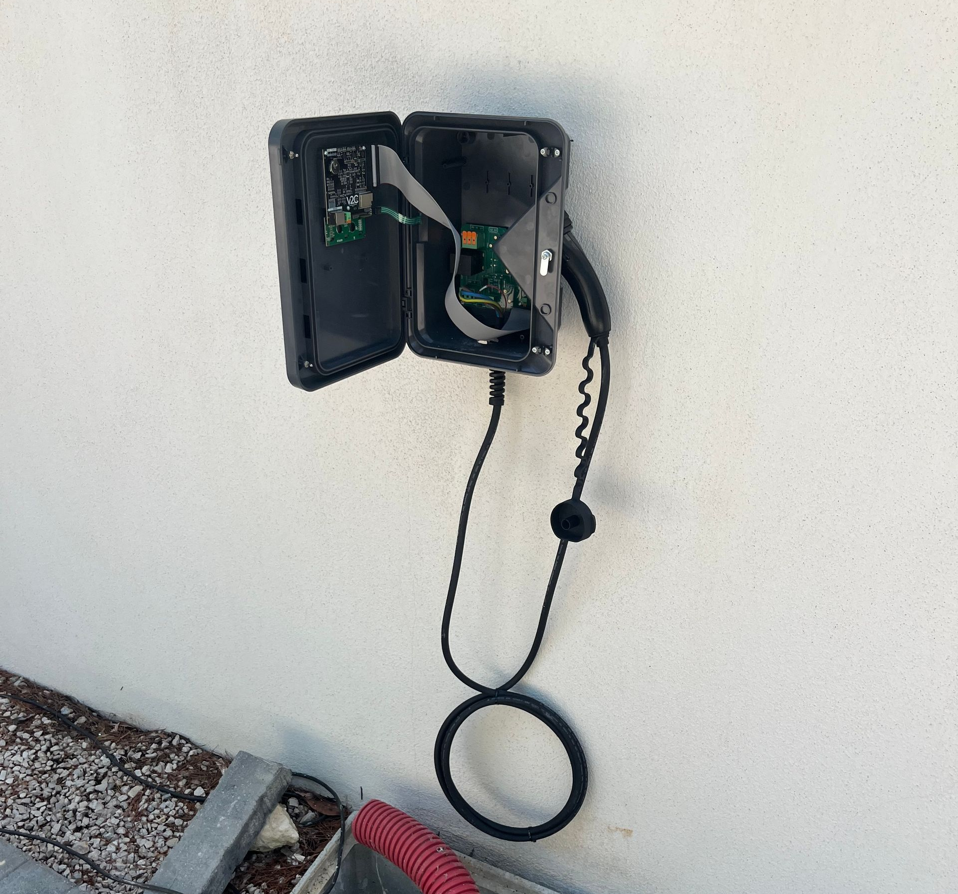 Chargeur de véhicule électrique noir endommagé, ouvert sur un mur blanc, câble suspendu.