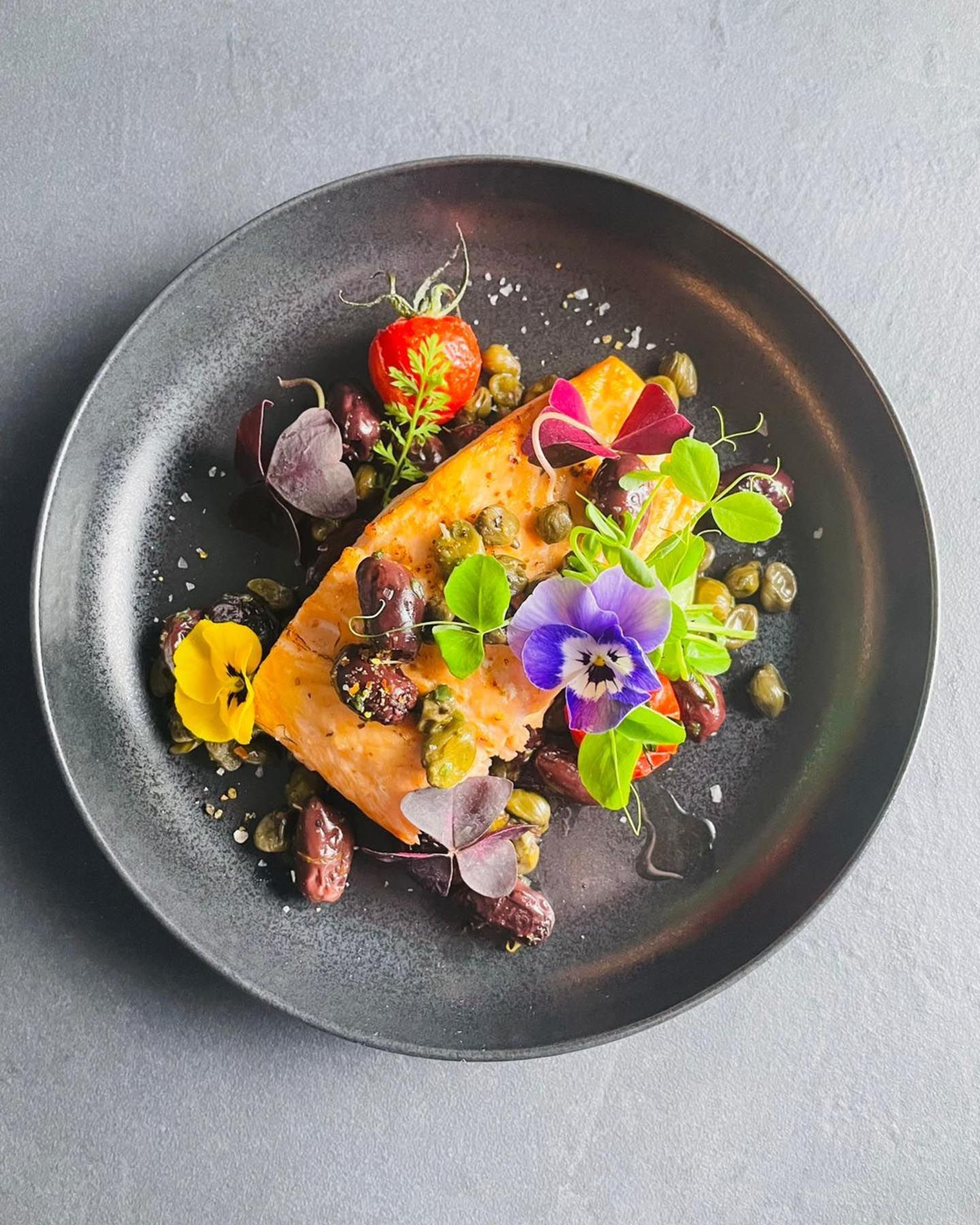 Lachs mit essbaren Blüten und Kräutern auf einem runden schwarzen Teller angerichtet