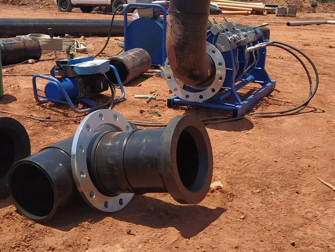 Codo de tubería industrial de HDPE con brida metálica, situado en una obra de construcción de tierra cerca de equipos de soldadura de tuberías.