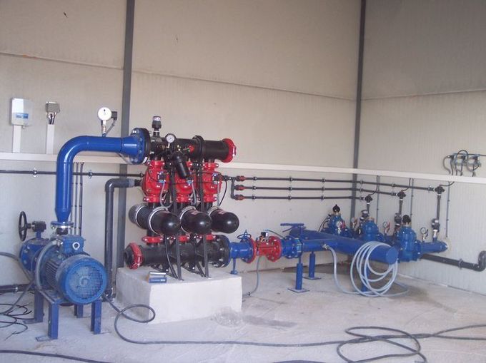 Un sistema industrial de filtración de agua con bombas azules, unidades de filtro negras y tuberías en una instalación interior blanca.