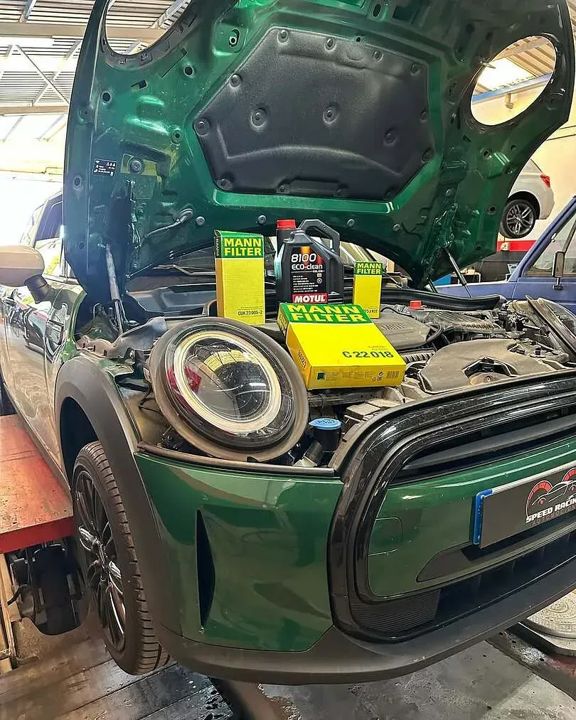 Un Mini Cooper verde está siendo reparado en un garaje con el capó abierto.