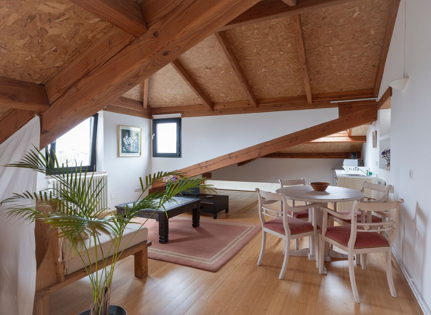 Sala de estar moderna en el ático con vigas de madera, sofá, mesa de centro, zona de comedor y grandes ventanales.