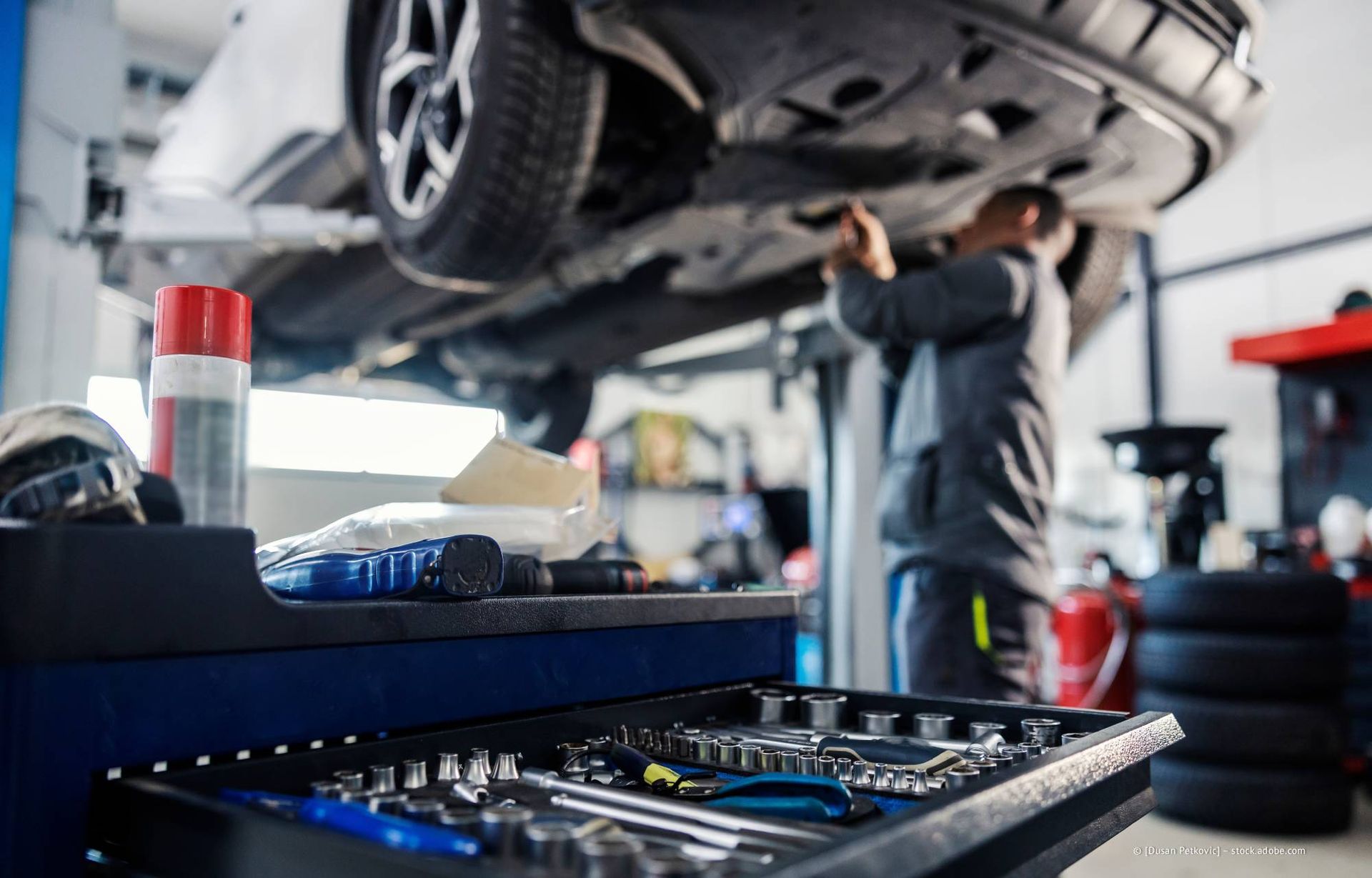 Ein Mechaniker arbeitet in einer Werkstatt unter einem auf einer Hebebühne befindlichen Auto. Im Vordergrund sind Werkzeuge in einem Tablett zu sehen.